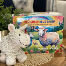 Load image into Gallery viewer, A Children's storybook by Maggie O'Hara -Red Sheep Blue Sheep – A Tale of Diversity and Acceptance" displayed on a modern coffee table, with a soft plush sheep toy, perfect for story time and teaching inclusion.