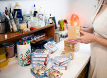 Load image into Gallery viewer, Maggie O'Hara holding a gift wrapped box of 'Faces of the Forest' spiritual affirmation cards stacked neatly on a shelf, designed to inspire kindness, creativity, and connection to nature.